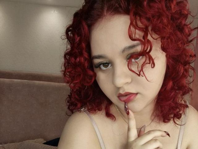 RosalieVittoria with curly red hair, fair skin, and subtle makeup, biting her red-painted fingernail, looking at the camera with a slightly pouty expression, wearing a light pink top.