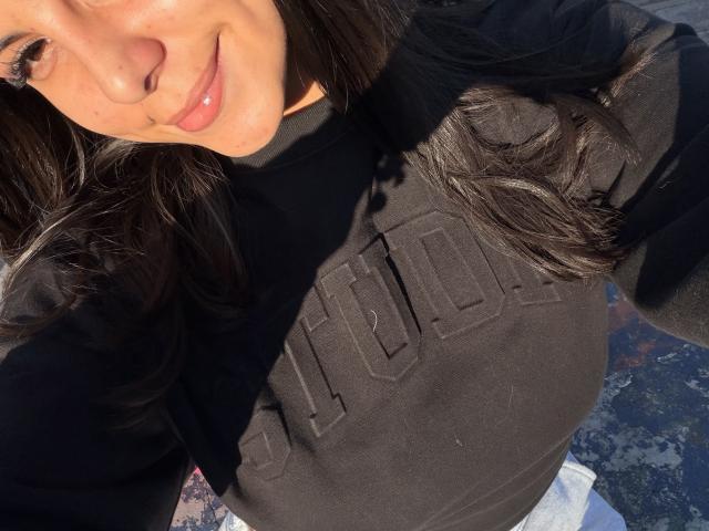 SarahPrincess with dark wavy hair, black shirt, and pink lips, smiling, showing her tits in a sunny selfie, shadow on her face.
