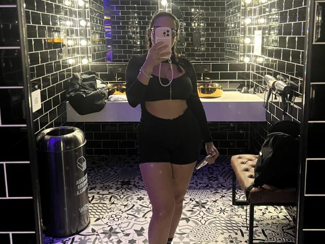 GalaMalkova with a slim, curvy body, wearing a black crop top and shorts, taking a mirror selfie in a black-tiled bathroom. Her ass is small, her tits are medium.