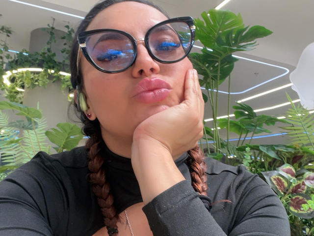 GalaMalkova, avec ses lèvres roses et pulpeuses, ses lunettes noires, ses cheveux bruns tressés, son pull noir à col roulé, une main sur sa joue, des écouteurs verts, et un regard confiant et adorable dans une pièce lumineuse..