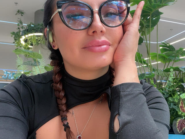 GalaMalkova with reflective glasses, pink glossy lips, black braided hair, black top, hand on cheek, necklace, and a cute face, showing a bit of her tits.