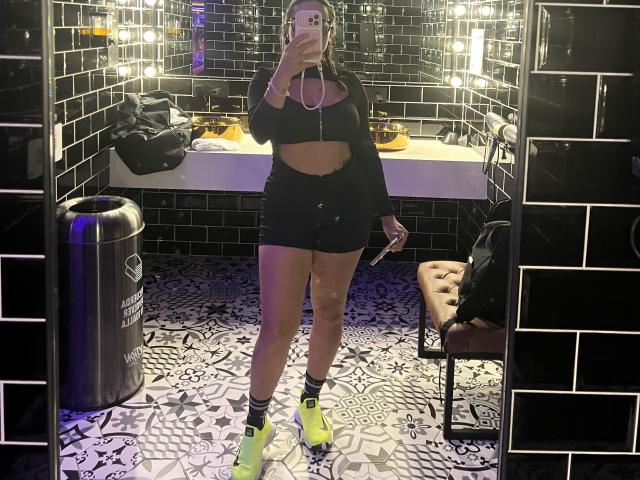 GalaMalkova with long brown hair, black crop top, black mini skirt, yellow sneakers, holding phone, standing in black-tiled bathroom, mirror reflection, trash can, brown tufted chair.