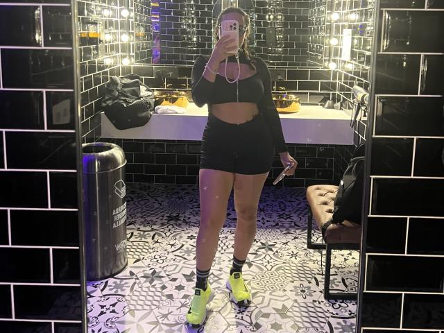 GalaMalkova with long brown hair, black crop top, black shorts, neon yellow sneakers, holding phone, mirror selfie, black tile walls, patterned floor, brown tufted chair, trash can.
