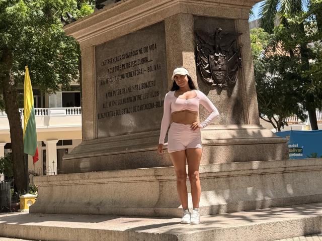 SarahPrincess standing in front of a stone monument, wearing a pink long-sleeve crop top, matching pink shorts, white sneakers, and a white cap, with long dark hair.