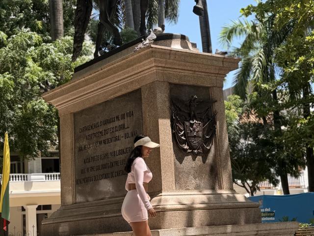 SarahPrincess with big ass, small white cap, pink long-sleeve and shorts, standing in front of a tall stone monument with a dark metal eagle sculpture on top.