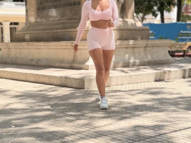 SarahPrincess with tan skin, pink long-sleeve crop top, pink shorts, white sneakers, walking, showing her ass, medium-sized boobs, and a curvy, fit body, in daylight.