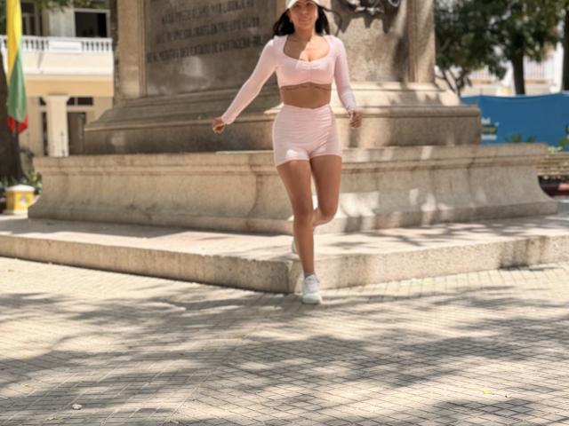 SarahPrincess with tanned skin, small tits, and a toned ass, wearing a pink long-sleeve crop top and matching pink shorts, running in a sunny urban area.