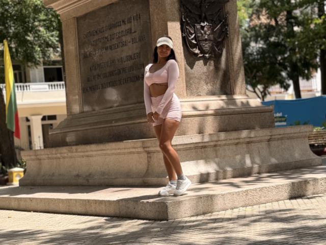 SarahPrincess stands in front of a stone monument, wearing a pink long-sleeve crop top and matching shorts, white sneakers, and a white hat, with her hands on her ass.