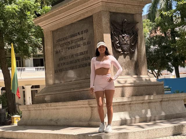 SarahPrincess stands in front of a stone monument, wearing a pink long-sleeve crop top and matching high-waisted shorts, white sneakers, and a white cap, with a serious expression.