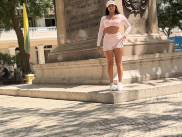 SarahPrincess stands with her ass in pink shorts, wearing a pink crop top, white sneakers, and a white cap, in front of a stone monument.