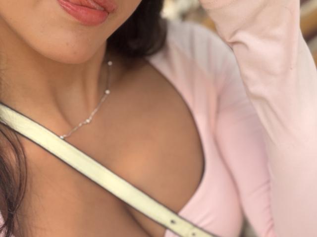 SarahPrincess's tits are visible in a pink top, with her tongue slightly out. She has a silver necklace and her shoulder is partially covered by a white strap.