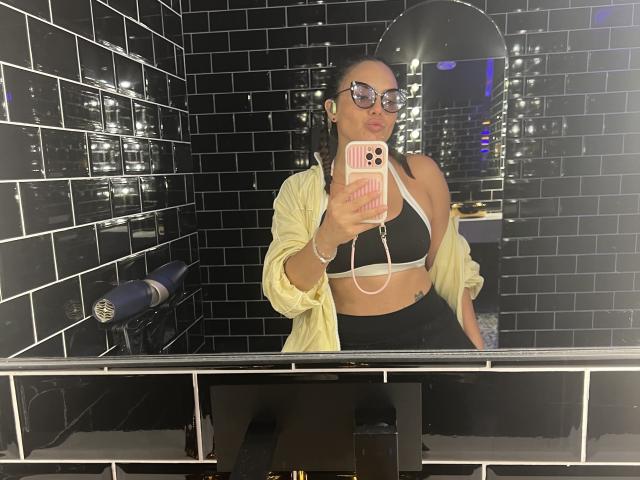 GalaMalkova with glasses, pink phone case, black bra, yellow shirt, black pants, holding phone in mirror, reflecting black tiled wall, mirror showing city lights. Boobs visible.