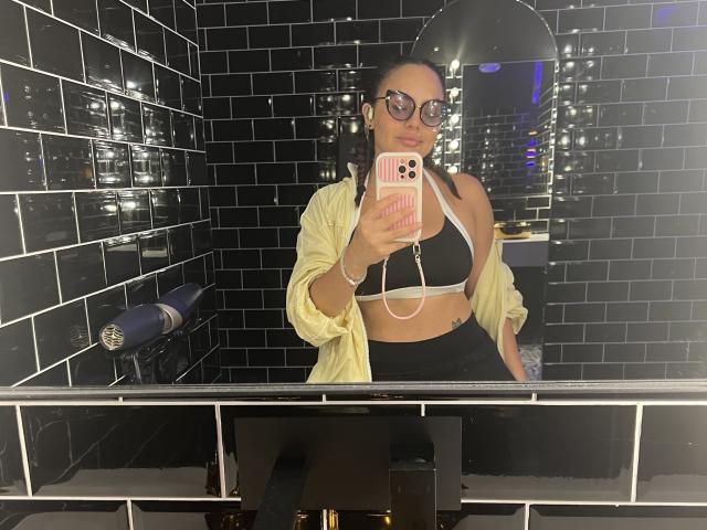 GalaMalkova with black bra, yellow shirt, black skirt, glasses, holding pink phone, mirror selfie in black-tiled bathroom. Boobs visible, white strap from bra, mirror reflection.