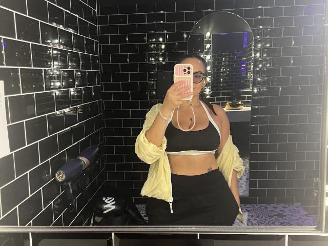 GalaMalkova with medium tits, wearing a black sports bra and black skirt, yellow shirt off, black tiled walls, mirror selfie, pink phone, glasses, mask, in a bathroom.
