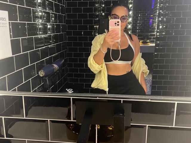 GalaMalkova with black sports bra, showing her boobs, wearing a yellow shirt off her shoulders, black pants, and glasses, taking a mirror selfie in a tiled bathroom.