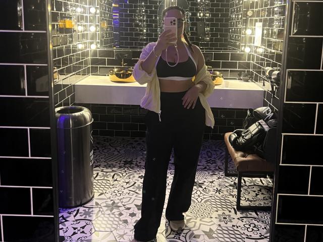 GalaMalkova with boobs in a sports bra, wearing black pants, a yellow shirt, and white sneakers, taking a mirror selfie in a tiled bathroom.