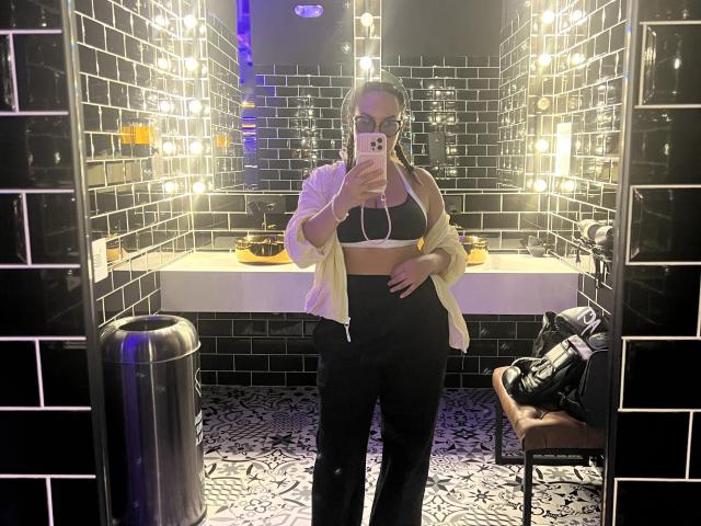 GalaMalkova with tits in a black sports bra, wearing black pants and a yellow jacket, taking a mirror selfie in a lit bathroom. Her hand is on her belly.