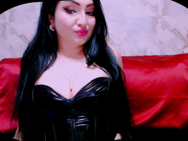 AlizeeChaillot with black shiny latex corset, large tits, long black hair, pink lipstick, gold necklace, pouty expression, red pillow behind her.