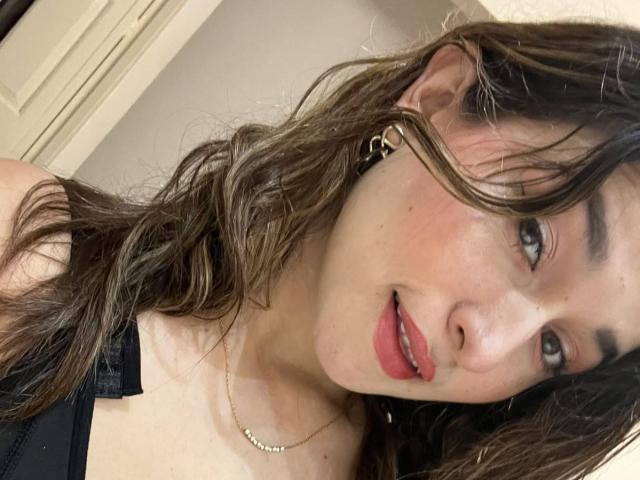 MilaGonz with wavy brown hair, red lips, and a gold necklace, showing her tits in a black bra, looking at the camera with a slight smile.