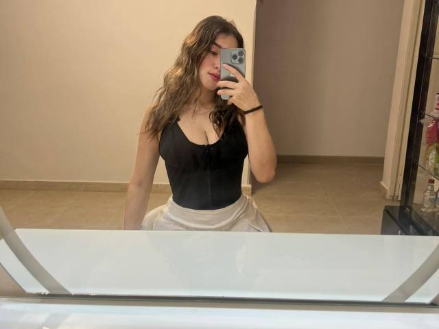 MilaGonz with wavy brown hair, wearing a black, low-cut, tight top, showing her big, round, juicy tits, and a white skirt, taking a mirror selfie with a smartphone.
