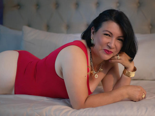 AmandaKohlerX, una donna asiatica formosa con capelli neri, sdraiata su un letto con un costume intero rosso, che mostra il sedere, sorridente con rossetto rosa, che indossa gioielli dorati..