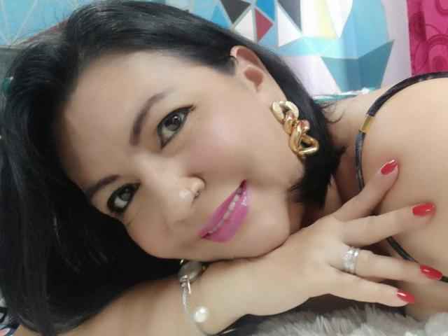 AmandaKohlerX with black hair, pink lipstick, red nails, gold earrings, and a nose ring, smiling at the camera, lying on a furry surface, showing her tits.