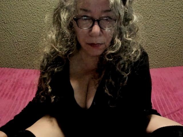 BeautyHairy with curly gray hair, glasses, and a black top, showing her tits in a low-cut shirt, sitting on a pink bed. She's looking at the camera with a slight smile.