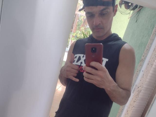 GuyFlexbi, com uma estrutura muscular, vestindo uma camiseta preta sem mangas e um boné preto, segurando um telefone vermelho, tirando uma selfie no espelho, exibindo seus braços tonificados..