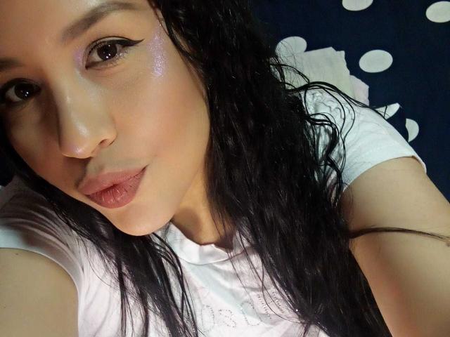 Il viso di MarySweett69, con la pelle rosa lucida, i capelli neri ondulati e l'eyeliner bianco, indossa una camicia bianca, è sdraiata, ha le labbra rosa chiare, guarda la fotocamera e sorride leggermente..