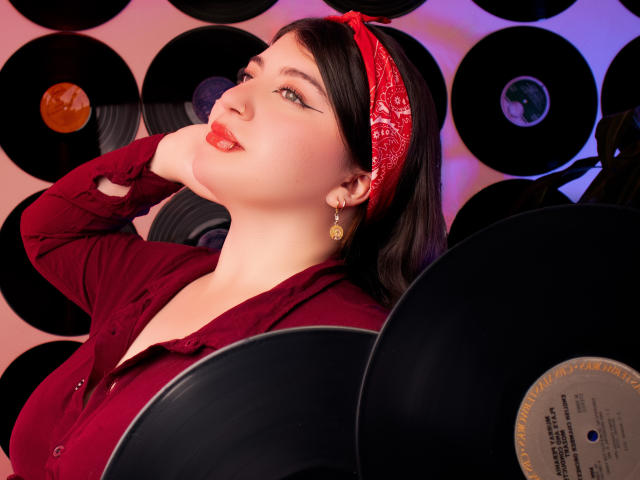 Cloe Larson, com um lenço vermelho na cabeça, camisa vermelha e brincos, segurando discos de vinil, tem a mão no pescoço, olhando para cima com batom vermelho, cabelo escuro e um rosto bonito..