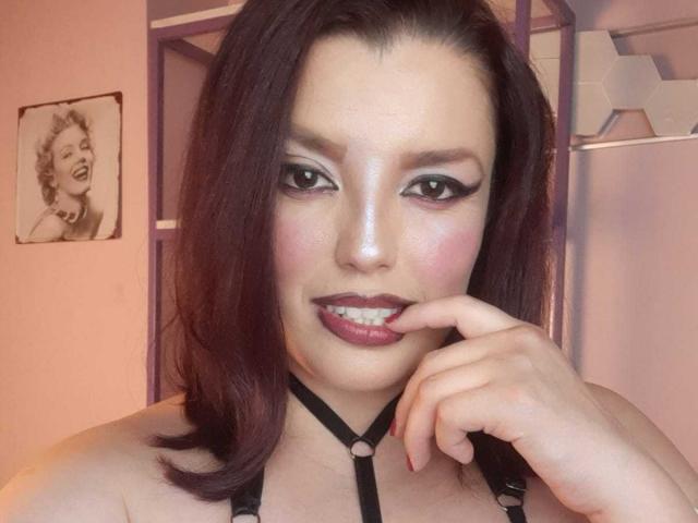 SachyMoon has dark purple hair, bold eyeliner, pink blush, and deep red lipstick. She's biting her finger, wearing a black harness, with a sexy, confident smile.