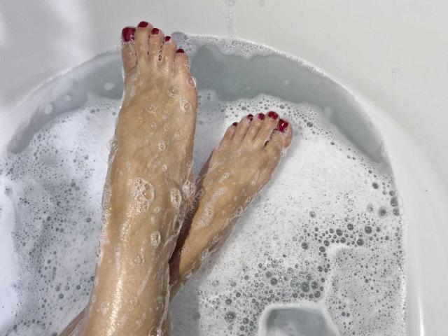KammyJoe's wet feet with red-painted toenails, soapy and bubbly, in a white bathtub. Water droplets on their skin, legs crossed at the ankles.