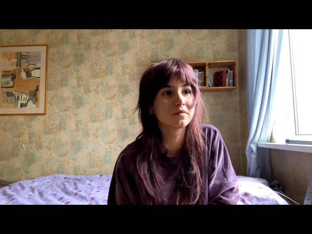 ShenikaTrucker, com cabelo roxo escuro, vestindo uma camisa roxa, sentada em uma cama com uma expressão séria, em um quarto com papel de parede verde claro estampado..