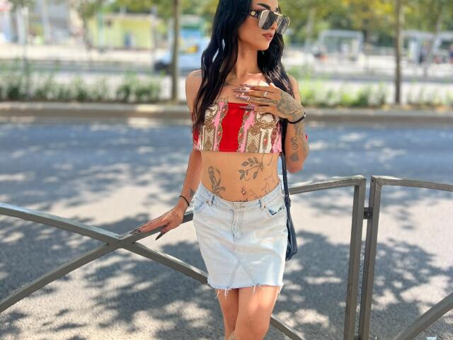 IrisIrs has tats, wears sunglasses, a red and gold crop top, and a frayed denim skirt, standing by a metal railing, showing her tits and a belly button.