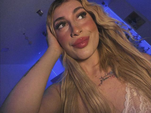 SammyViolet, com cabelo loiro, lábios vermelhos e um piercing no nariz, tocando a cabeça, vestindo uma blusa de renda, mostrando os seios e com uma tatuagem no peito..
