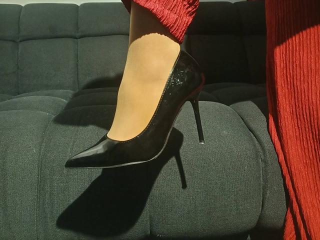 MissEellen's shiny black high heel with a pointy toe is perched on a dark green couch, casting a shadow. She's wearing red, textured pants.