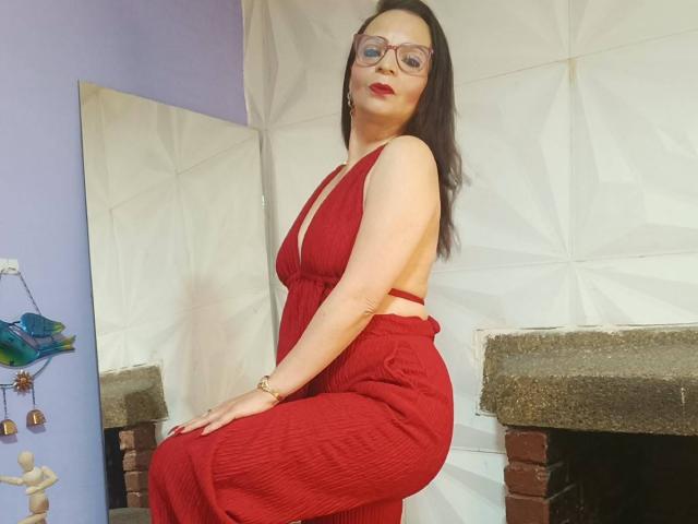 MissEellen with red dress, showing her ass, glasses, and red lipstick, sitting on brick fireplace, hand on knee, white geometric wall in background, purple wall to the left.