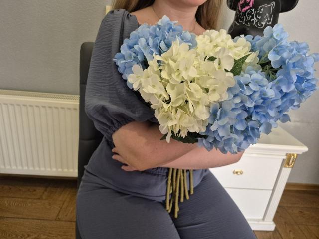 Meeranda with blue and white hydrangeas, wearing a gray dress, sitting on a chair, holding the bouquet in front of her, wooden floor, white dresser, and radiator.