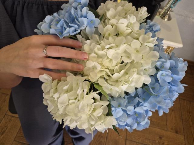 Meeranda's hand with a ring holds a bouquet of white and blue hydrangea flowers, her fingers gently touching the petals.