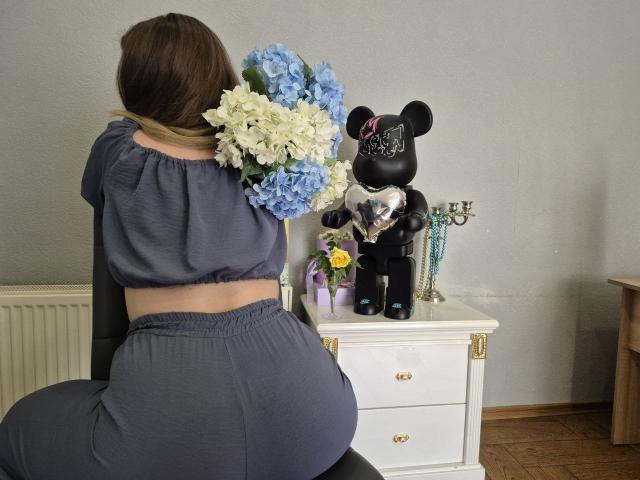 Meeranda with brown hair, wearing a blue crop top and matching pants, sits on a chair, facing a white dresser with a black Mickey doll and blue-white flowers.