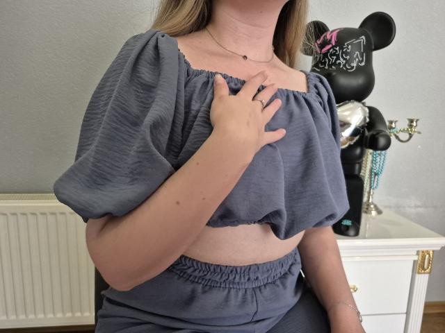 Meeranda's hand touches her gray, puffy-sleeve, cropped top, revealing her bare, soft tits. She's wearing matching gray pants, standing near a black Minnie Mouse statue.