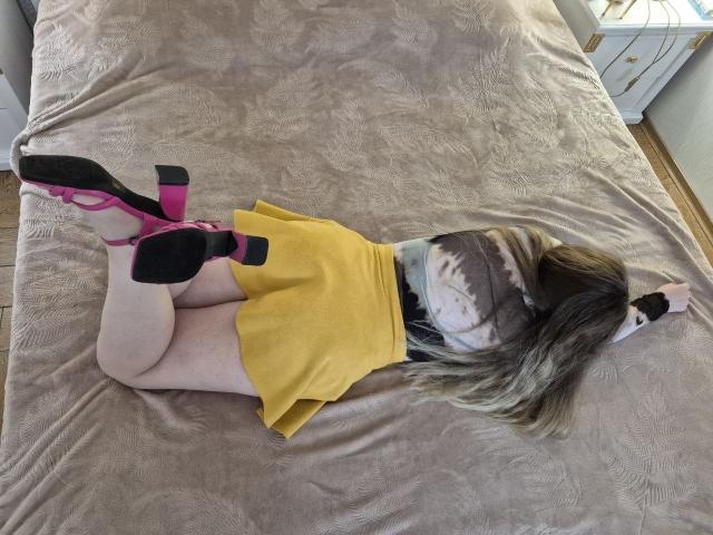 Meeranda, laying on a bed with her ass up, wearing a yellow skirt, pink high heels, and black gloves, has long blonde hair and pale skin.
