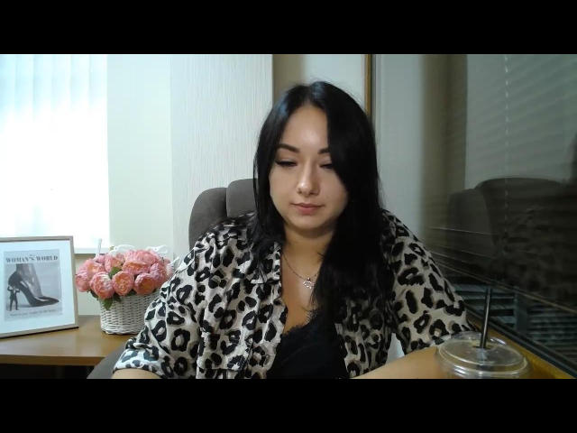 AdriannaGoldis with long black hair, wearing a leopard print robe, sitting at a desk, looking down, hands in front, black top, pink roses, framed photo, and glass cup.