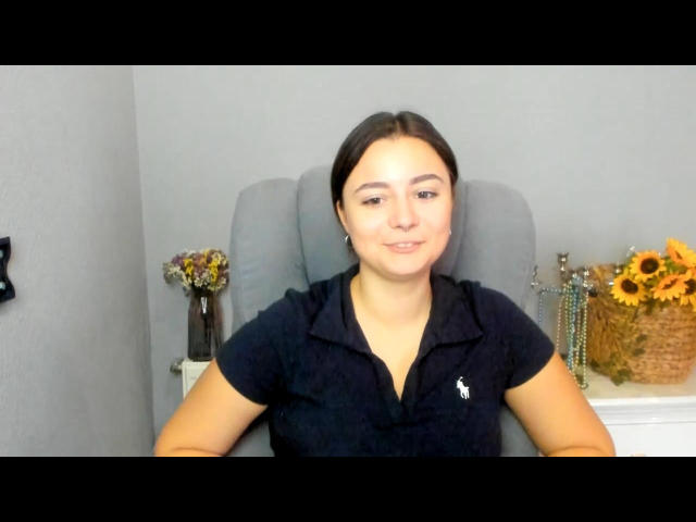 SofiSolty with brown hair, black polo shirt, sitting in grey chair, smiling, small tits, light skin, arms resting on chair, floral vase and sunflowers on right.