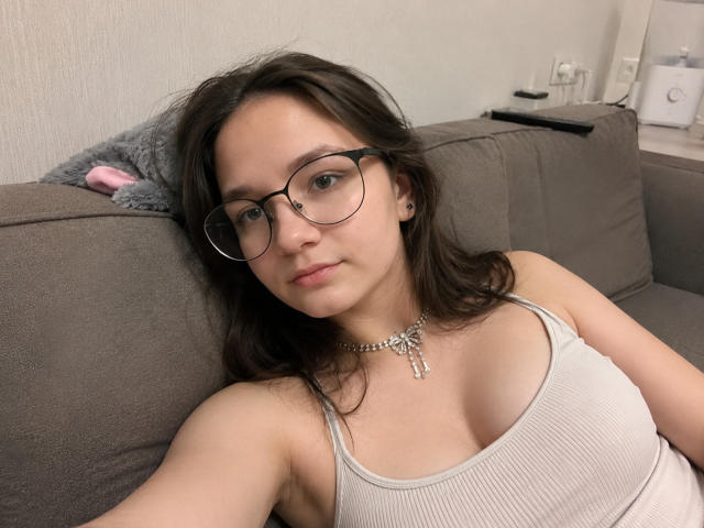 OdeliaHolen with glasses, light skin, and brown hair, wearing a ribbed tank top, showing her small tits, lying on a gray couch, necklace, and a neutral expression.