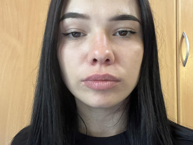 ElvaPaup hat schwarzes, glattes Haar, braune Augen mit schwarzem Eyeliner und rosafarbene Lippen. Sie blickt neutral in die Kamera und trägt ein schwarzes Hemd..