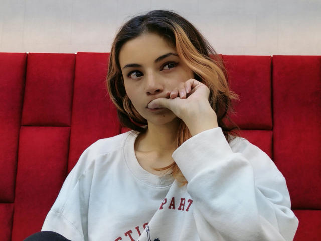 Amuneet with wavy brown and orange hair, wearing a white sweatshirt, resting her chin on her hand, looking at the camera with a slight pout, sitting on a red cushion.
