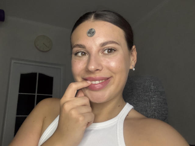 BlackFatal heeft een kleine blauwe cirkel op haar voorhoofd, ze glimlacht met roze lippen, bruine ogen, en een vinger die haar kin aanraakt. Ze draagt een witte tanktop en oorbellen..