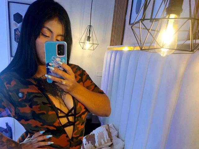 AngelaJoness with dark hair, wearing a camo dress with black lace-up front, holding a blue phone, showing tattoos, and taking a mirror selfie, with a light bulb above.