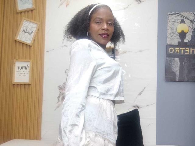 SarahiPusey with curly black hair, wearing a shiny white satin top and skirt, dark purple lipstick, white headband, standing with a black chair behind her.